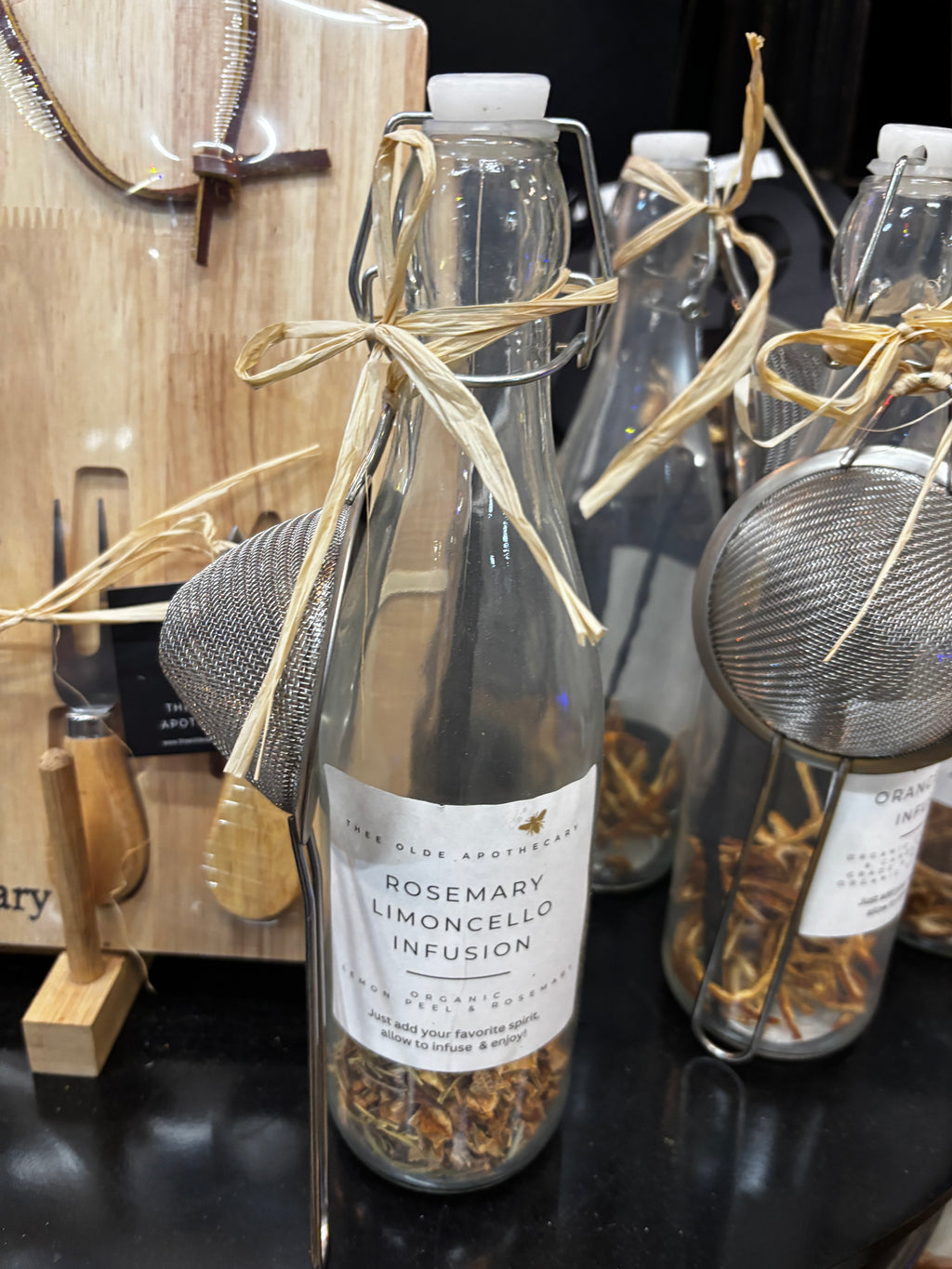 A clear glass decanter with a flip-top cap and a attached a beige ribbon, adorned with a label reading 'ROSEMARY LIMONCELLO INFUSION', along with a strainer and spices visible inside.