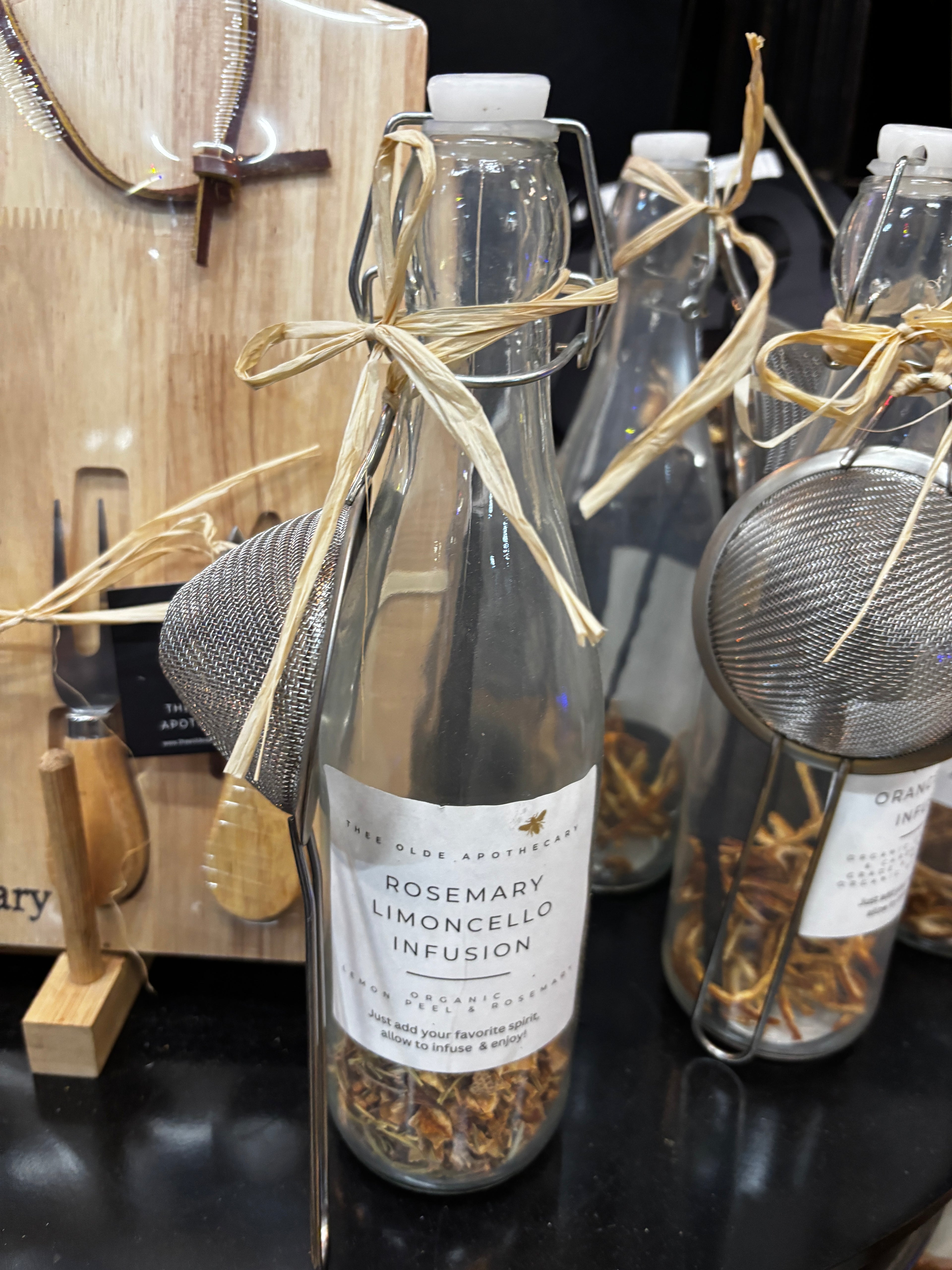 A clear glass decanter with a flip-top cap and a attached a beige ribbon, adorned with a label reading 'ROSEMARY LIMONCELLO INFUSION', along with a strainer and spices visible inside.