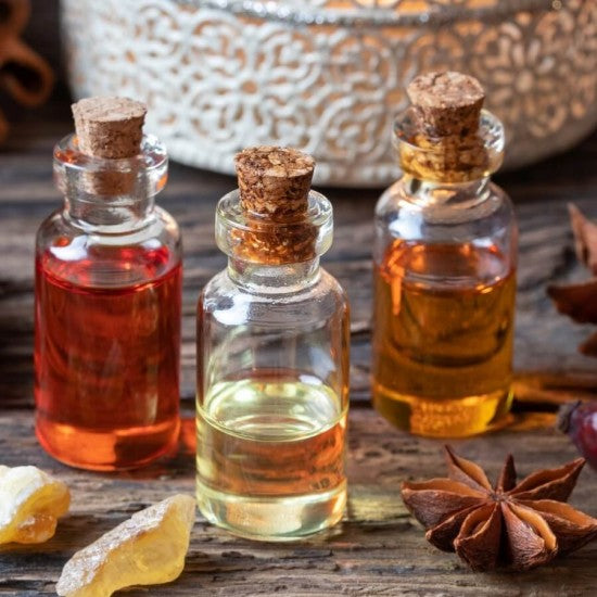 Three small glass bottles with cork stoppers, containing different colored liquids, likely to be essential oils. The bottles are displayed on a wooden surface with cork stoppers, lemon, and other aromatic botanicals scattered around.