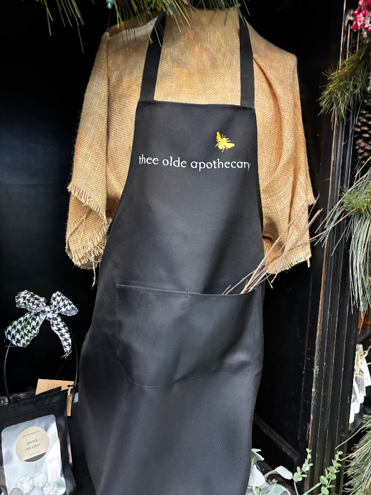 Black apron with 'thee olde apothecary' logo on a stand with decorative items.